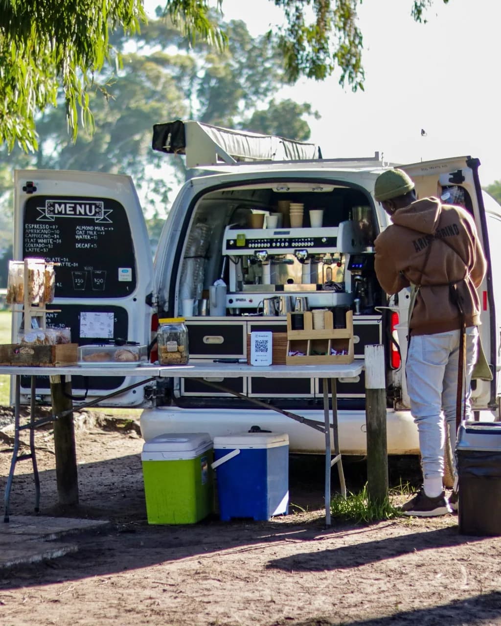 The open Arise & Grind coffee van beneath trees, with a barista preparing coffee beside the espresso machine.